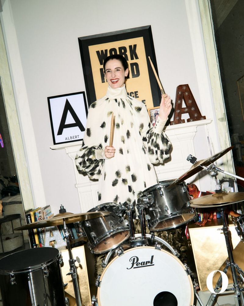 A woman in a patterned dress plays drums indoors, holding drumsticks with framed art and a Pearl drum set prominently displayed.