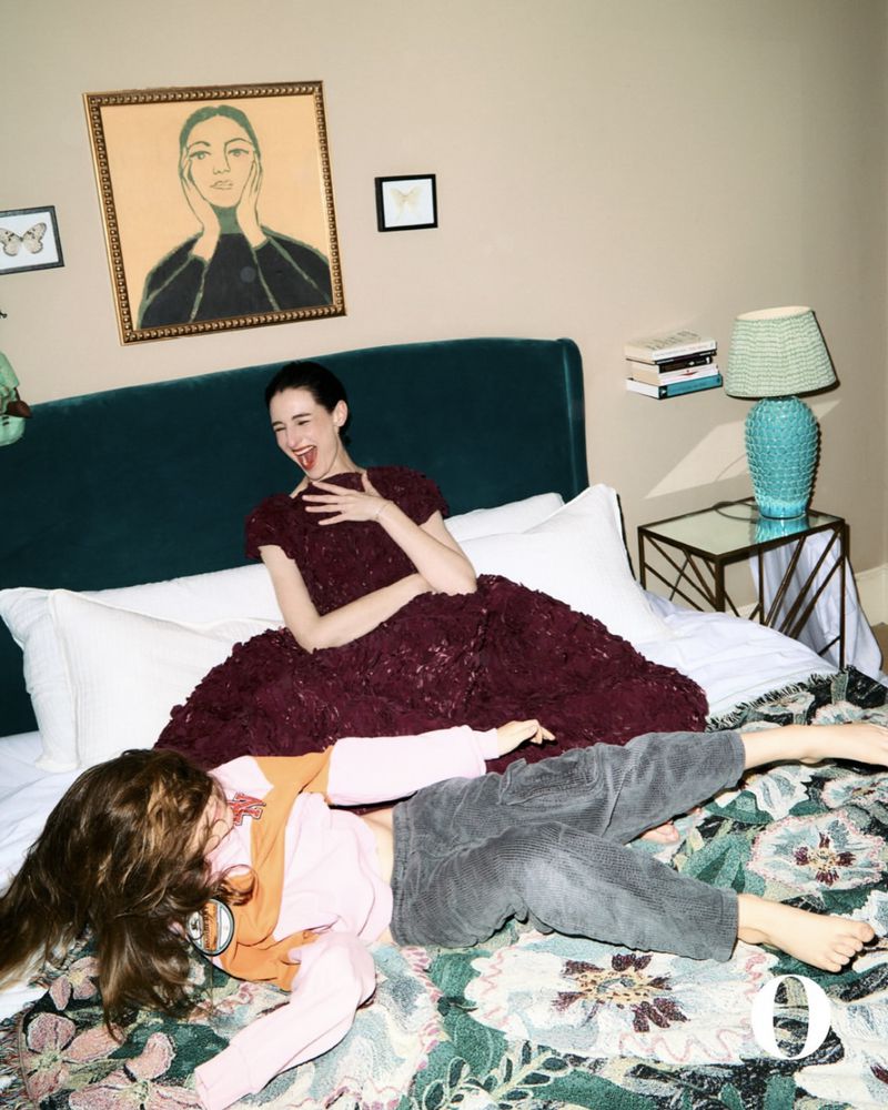 Two women on a floral bedspread; one woman in a formal dress sits up, the other in casual clothes lies at the foot of the bed.
