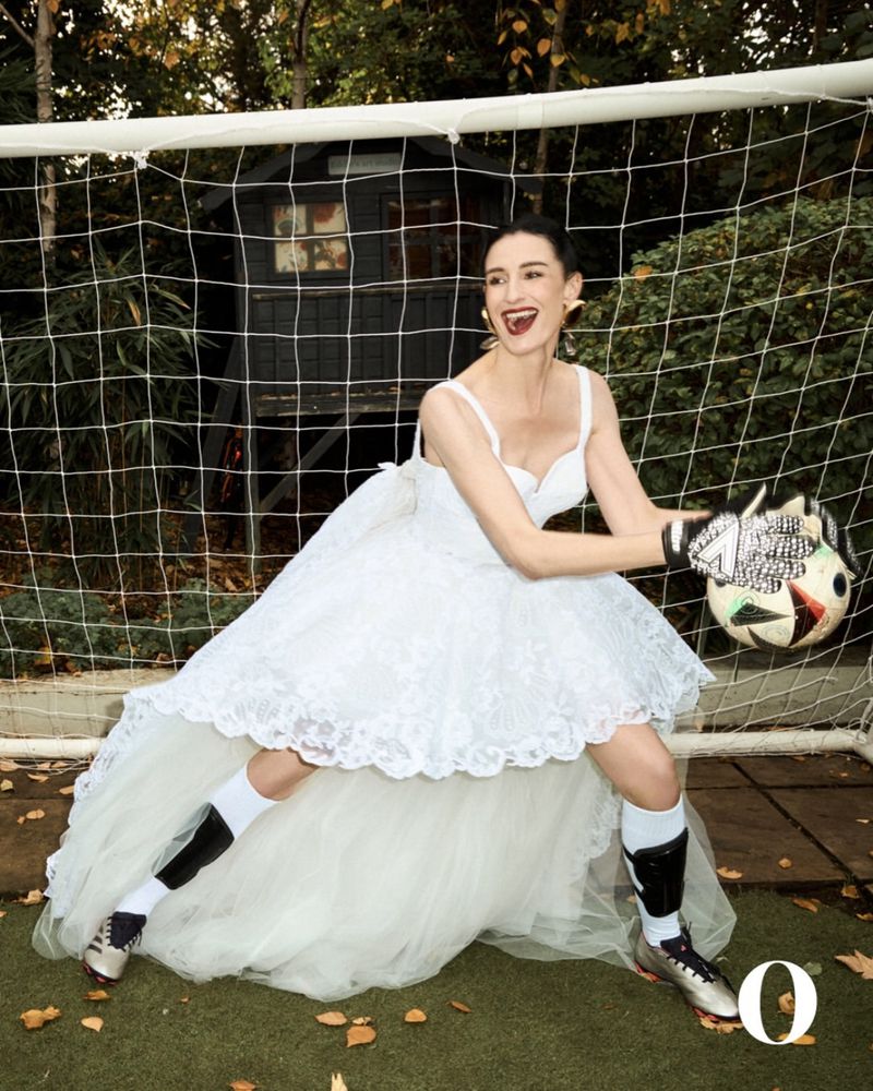A woman in a wedding dress, goalie gloves, and soccer socks poses near a soccer goal net outdoors on a grass field, holding a soccer ball.