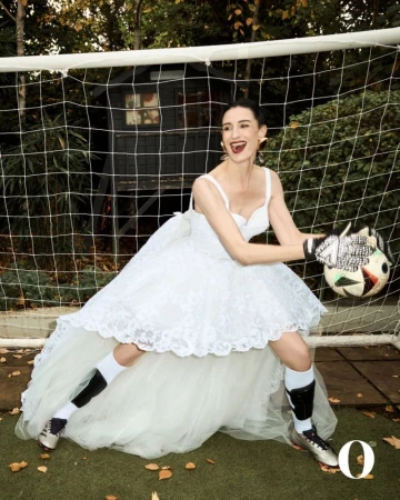 A woman in a wedding dress, goalie gloves, and soccer socks poses near a soccer goal net outdoors on a grass field, holding a soccer ball.