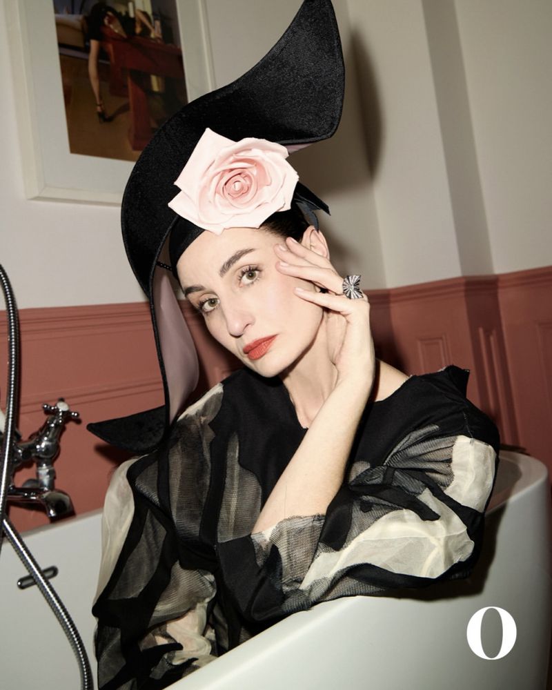 Woman in black hat with rose, posing in bathtub. Close-up captures her stylish look with vintage-style jewelry and modern fashion.