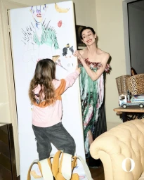 Model Erin O'Connor standing beside a large vertical drawing board being decorated by a child in a pink hoodie