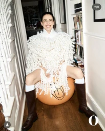 Model Erin O'Connor laughing while seated on an orange space hopper in a hallway wearing a textured white knit and tall brown boots.