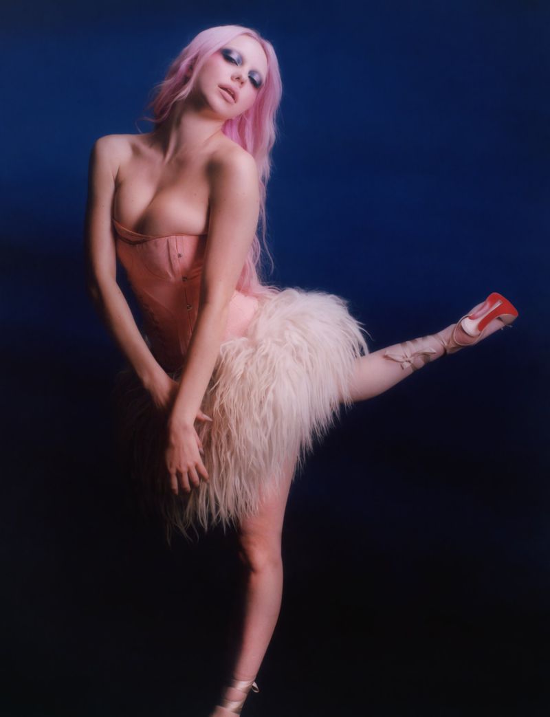 Elegant woman with pink hair poses in a pink corset dress and fluffy skirt in a studio, striking a playful ballerina pose.
