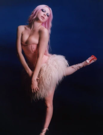 Sienna Spiro with pink hair wearing a blush corset and feathered skirt striking a ballet pose against a navy blue backdrop.