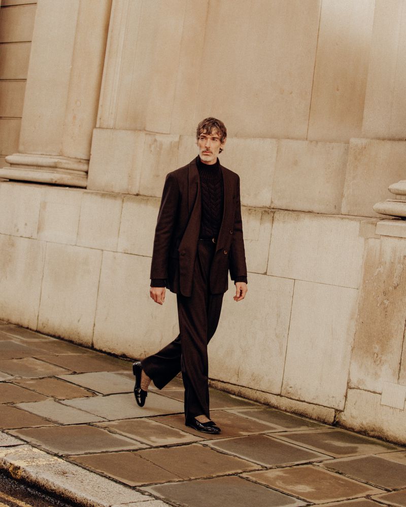 A confident man in a suit strides along a stone sidewalk next to a textured wall, capturing a blend of fashion and urban sophistication.