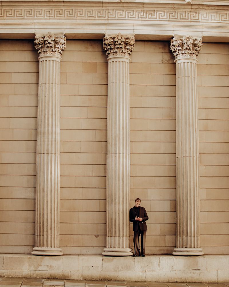 A man leans against a large column with decorative corinthian style capitals, wearing a suit in front of a neutral colored stone wall.