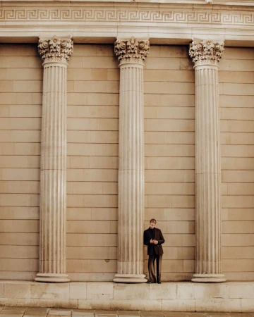 A man leans against a large column with decorative corinthian style capitals, wearing a suit in front of a neutral colored stone wall.