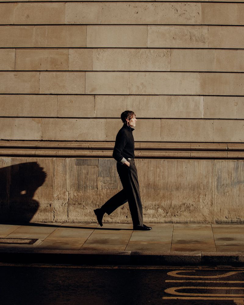Adult male walking on a sidewalk in front of a textured tan stone wall, wearing a black turtleneck, grey trousers, and black shoes.