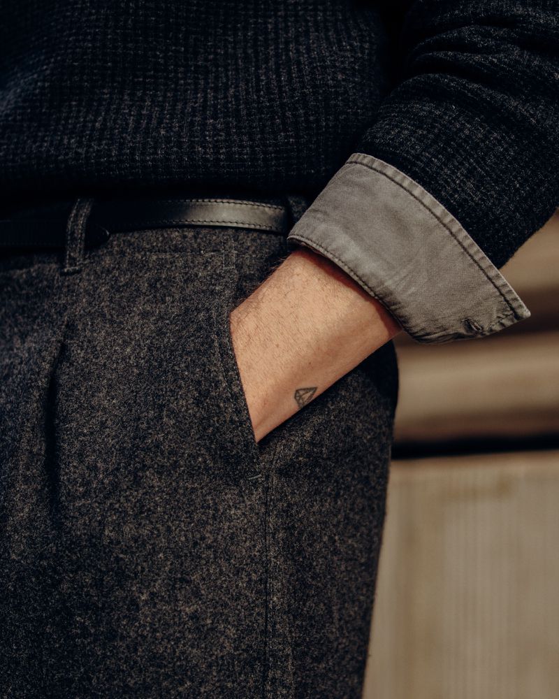 Close-up of a man's torso and arm, featuring a dark sweater, grey trousers with a belt, and a small tattoo on his hand.