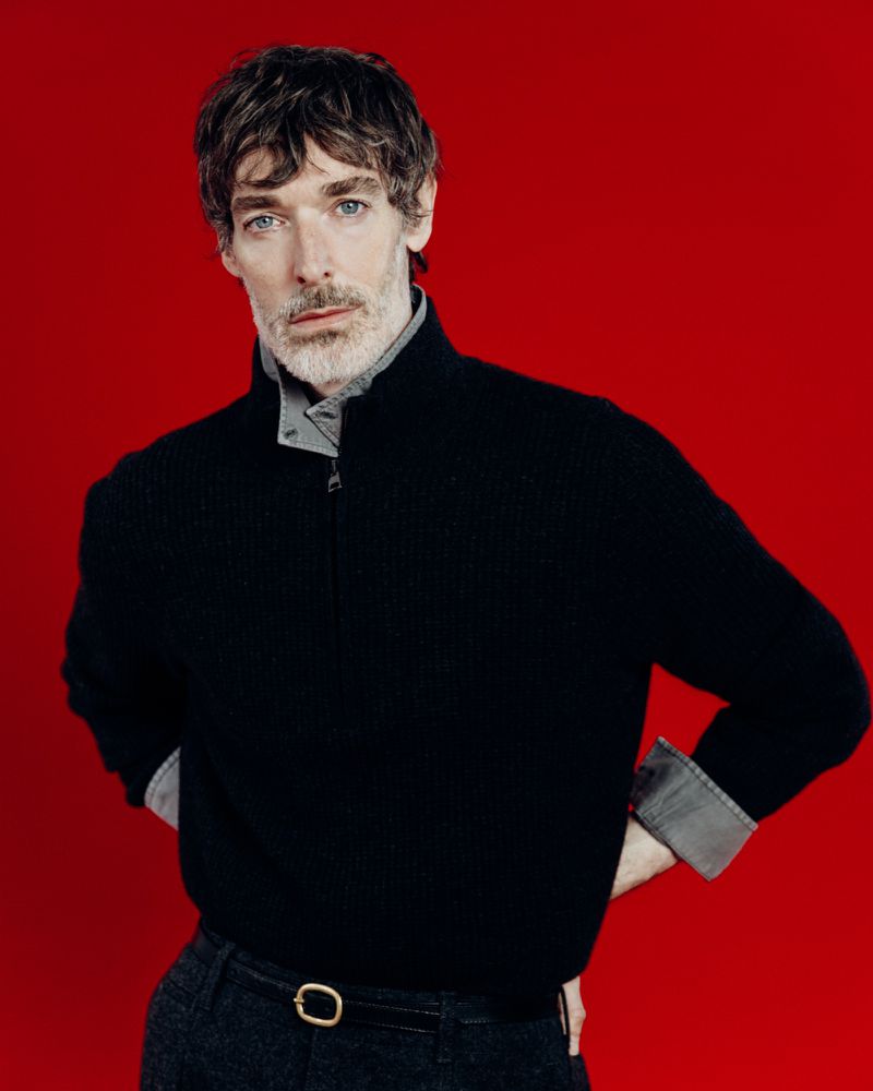 Portrait of a middle-aged man with wavy brown hair against a solid red background, wearing a black sweater and grey shirt.