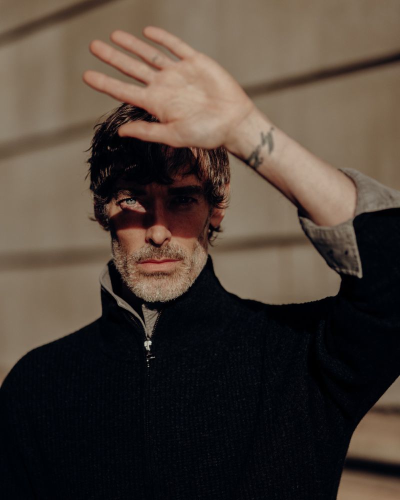 Portrait of adult male with hand raised shielding face from sunlight. He wears a black jacket, and has a light beard and blue eyes.