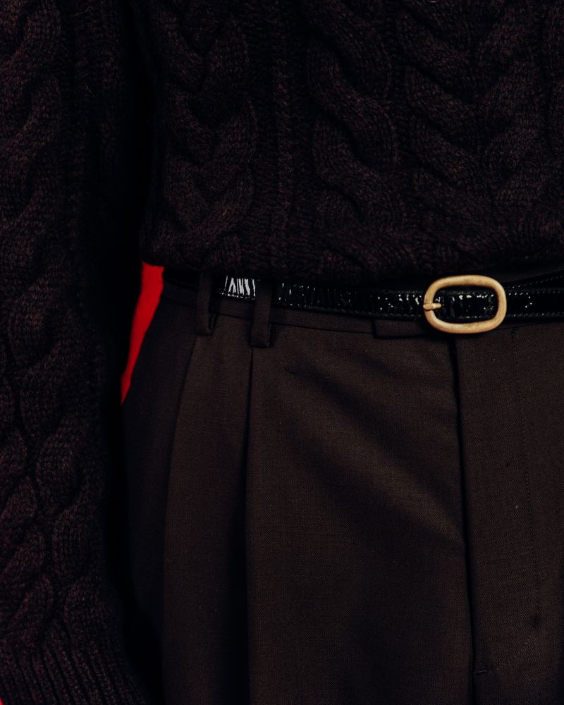 Close-up of a dark brown knit sweater paired with dark brown pleated pants and a thin black patent leather belt with a gold buckle.