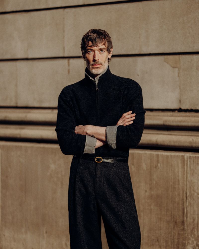 Portrait of a stylish adult man in a black cardigan and grey shirt with arms crossed, standing against a textured wall in sunlight.