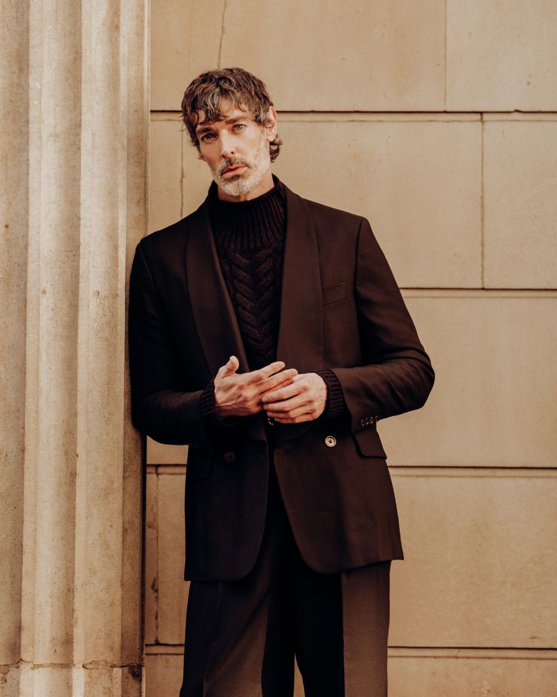 Portrait of an adult male in a dark suit and turtleneck sweater leaning against a concrete pillar, looking at the camera.