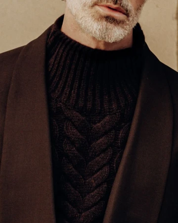 Close-up of a man with salt and pepper beard wearing a brown cable knit sweater and brown wool coat against a tan background.