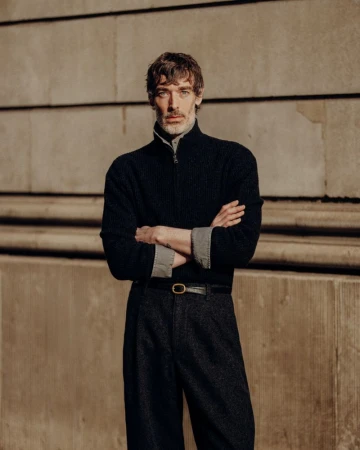 Model Richard Biedul poses with crossed arms wearing a dark zip knit sweater and pleated trousers against a warm stone wall.