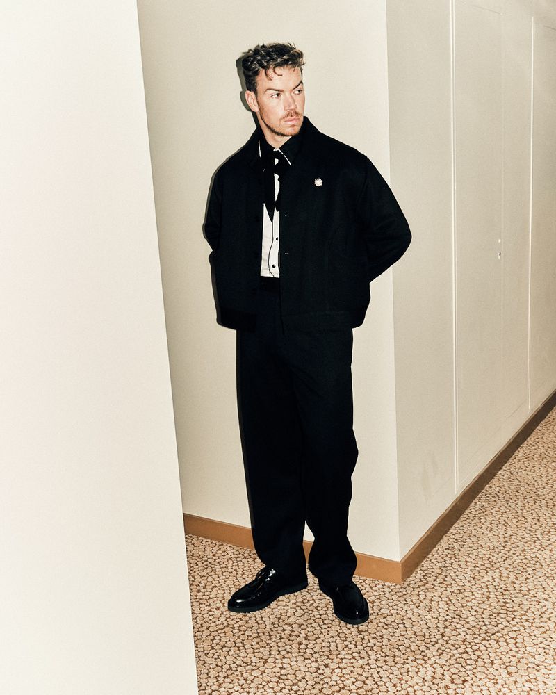 Stylish man in a black suit and white shirt posing indoors with a confident expression in a full-length portrait.