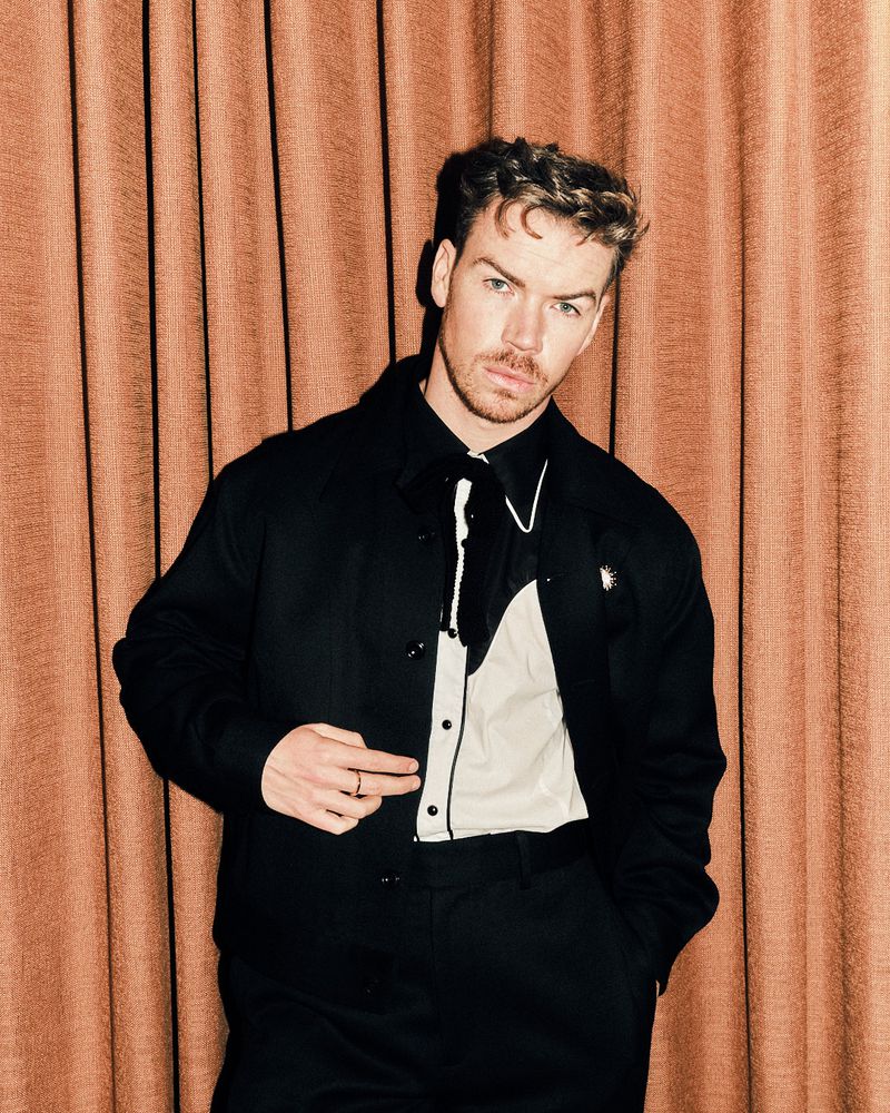 Portrait of a stylish adult male in a black suit and white Western-style shirt posing in front of a textured, peach-colored curtain.