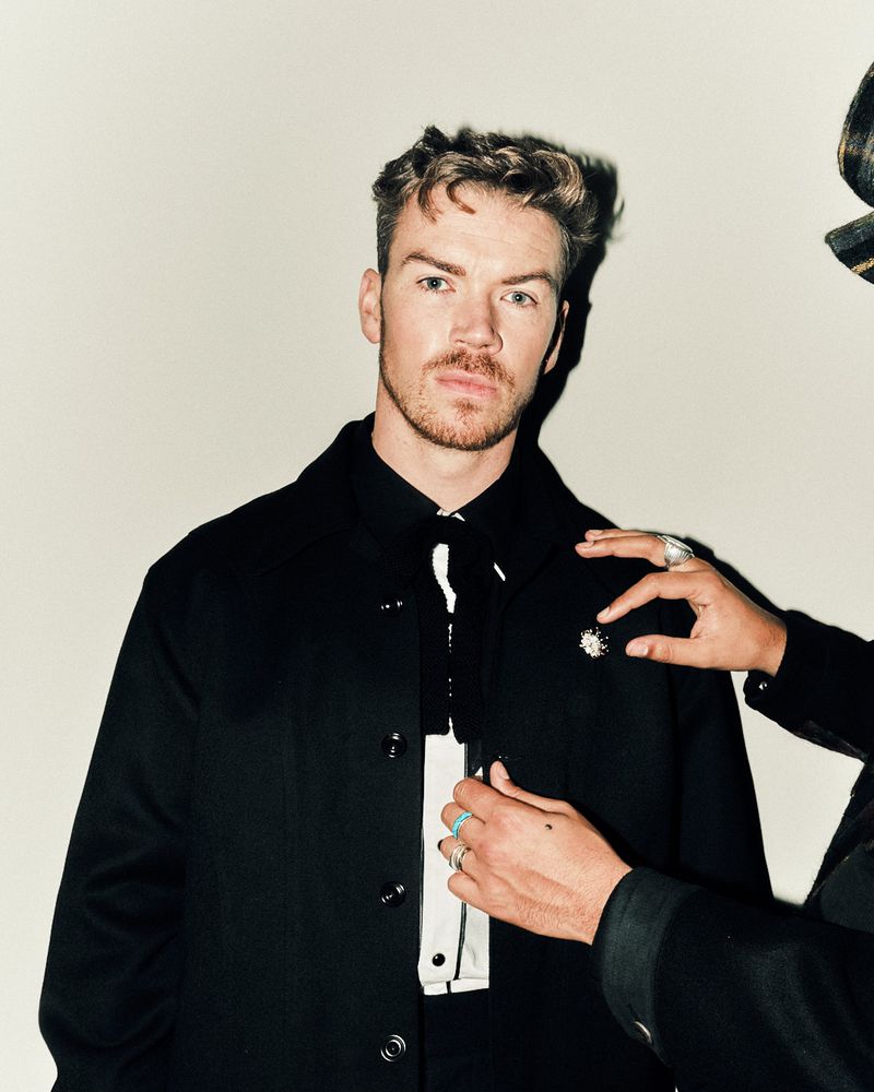 Portrait of a fair-skinned man in a black coat and black and white shirt being dressed by a person wearing rings and a hat.