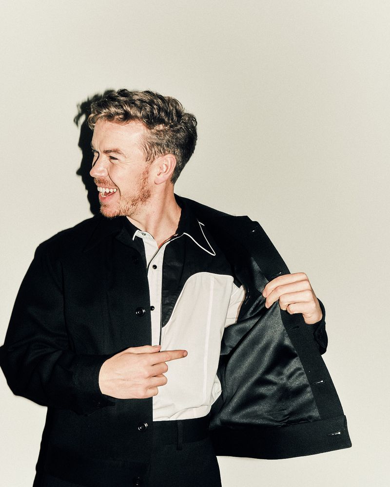 Portrait of a happy adult male with light brown hair wearing a black jacket and white shirt, pointing to the inside of his jacket.