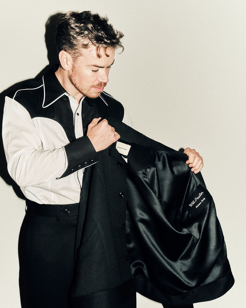 Portrait of a man in a black and white Western-style shirt, holding a black jacket, posing in front of an off-white backdrop.
