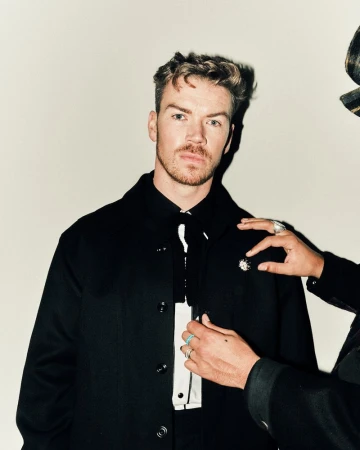 Will Poulter wearing a black jacket and white shirt being adjusted by another person against a plain wall.