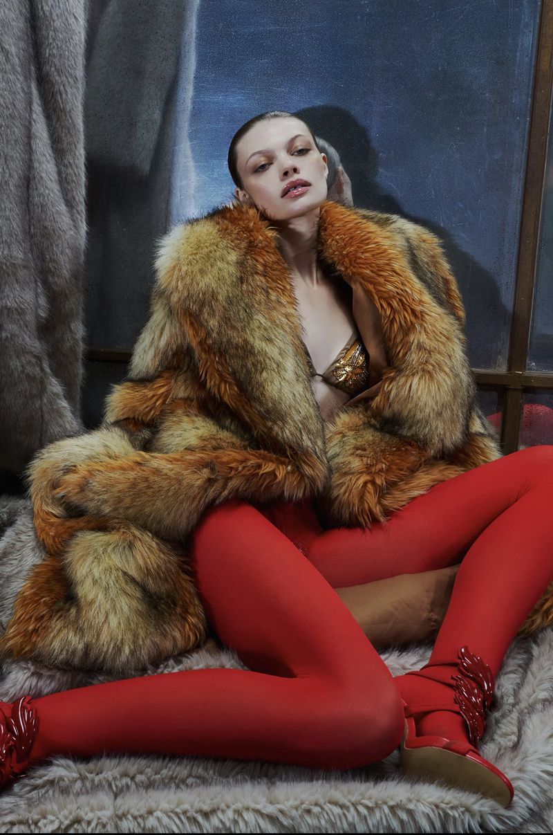 A fair-skinned fashion model poses in a fur coat and red tights against a neutral studio backdrop, showcasing a high-fashion editorial style.