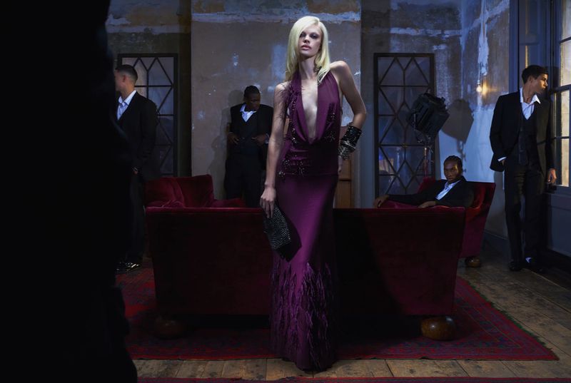 A blonde female model in a low-cut gown poses in an indoor environment surrounded by men in suits and a red velvet couch.