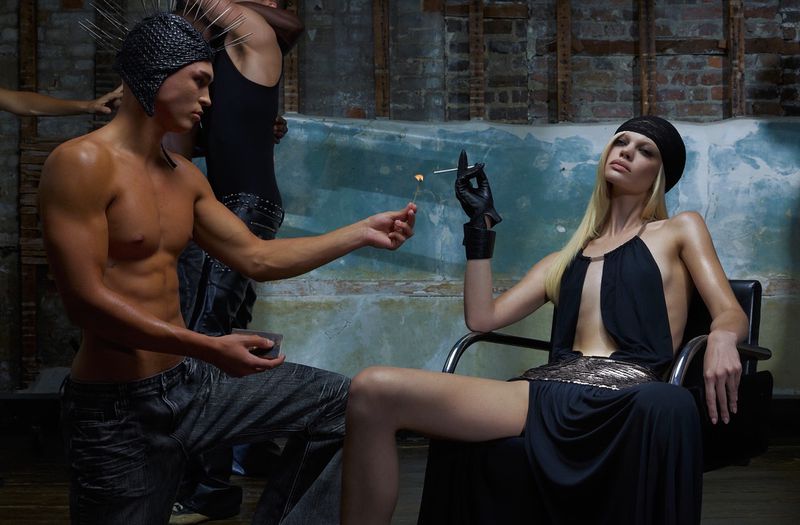 Fashion photo of a man lighting a match for a woman in a dress and headscarf in an indoor studio with brick walls and dark tones.