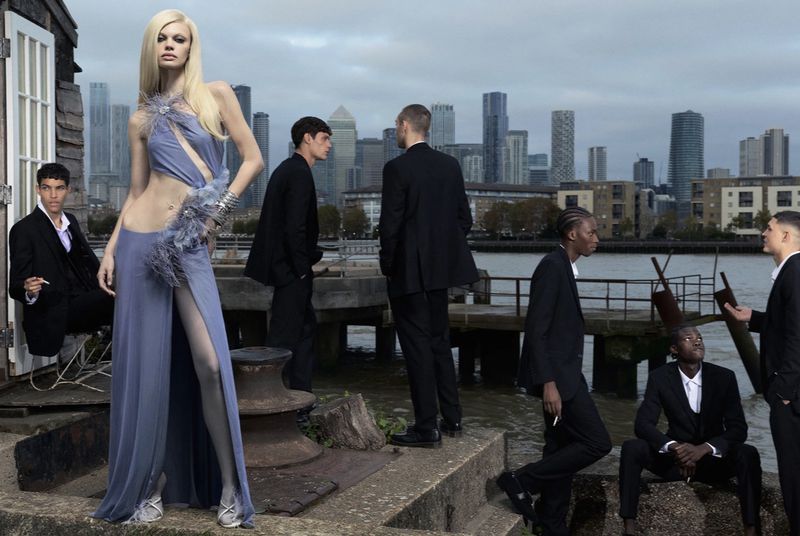 Fashion editorial shot with a fair-skinned woman in a lilac gown with feather details, posed with a group of men in dark suits by the water.
