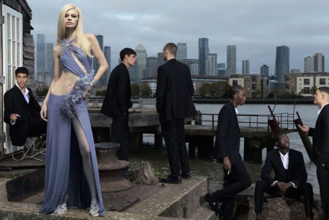 Model Lexie Tapper wearing a pale blue cutout feathered gown surrounded by men in black suits along an urban waterfront.