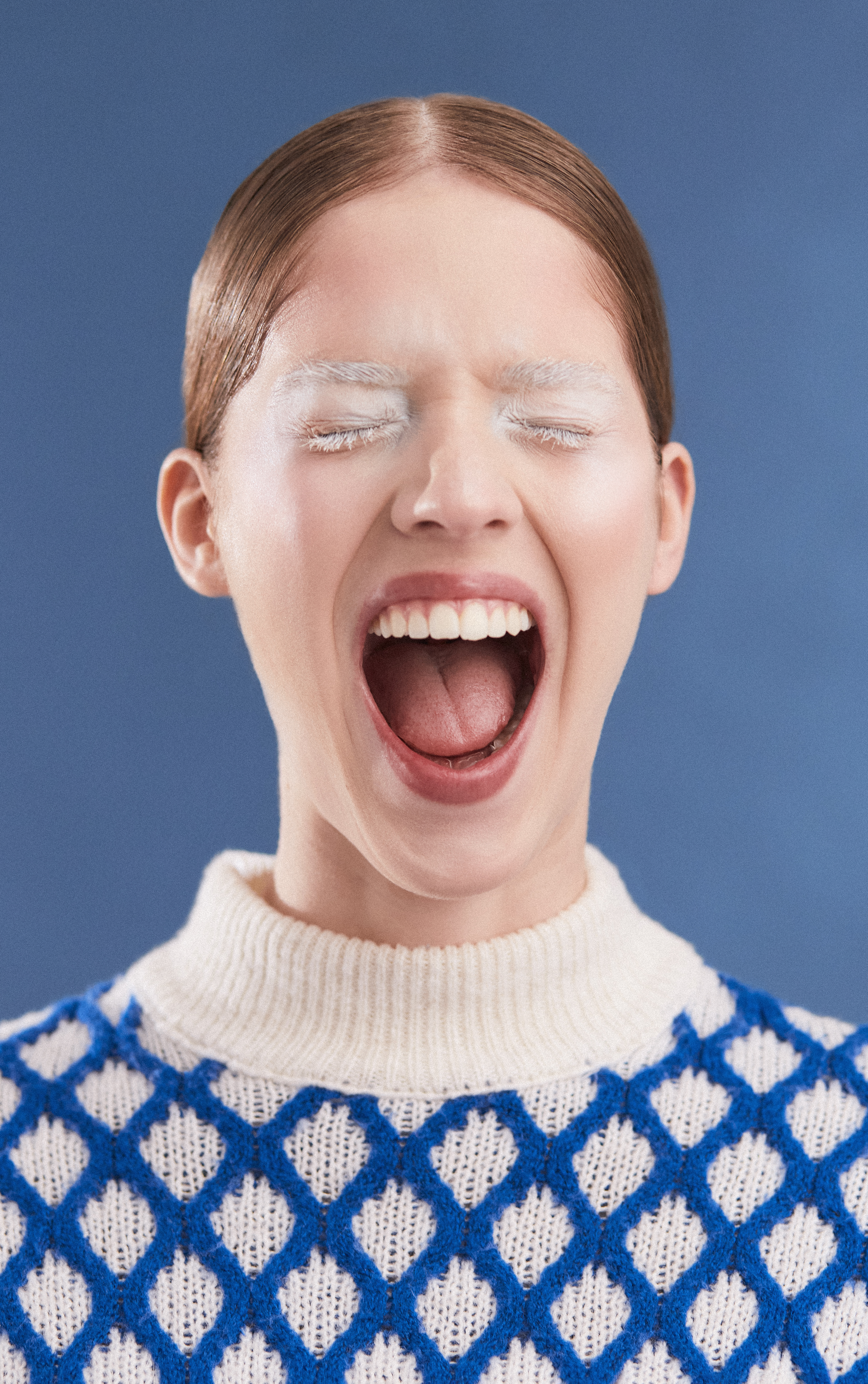 Close-up studio shot of a white adult female model with a wide-open mouth and closed eyes, wearing a blue-and-white sweater, on a blue background.