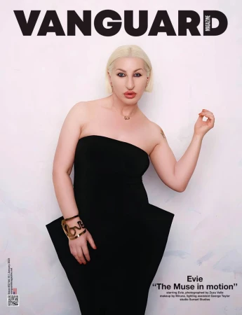 Blonde woman wearing a strapless black peplum dress and gold cuff bracelet posing against a textured white wall.