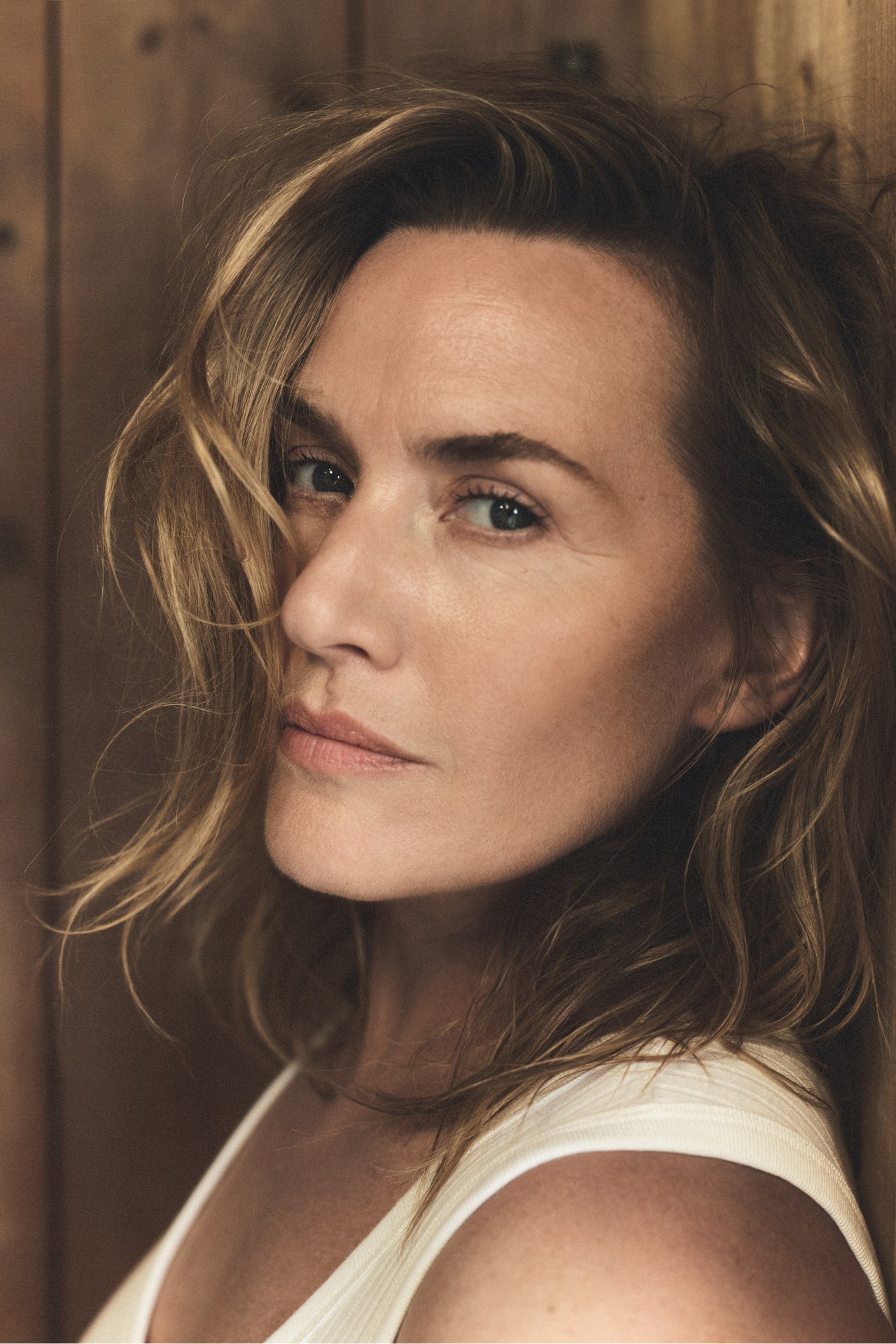 Portrait of actress Kate Winslet wearing a white top against a rustic wooden wall background.