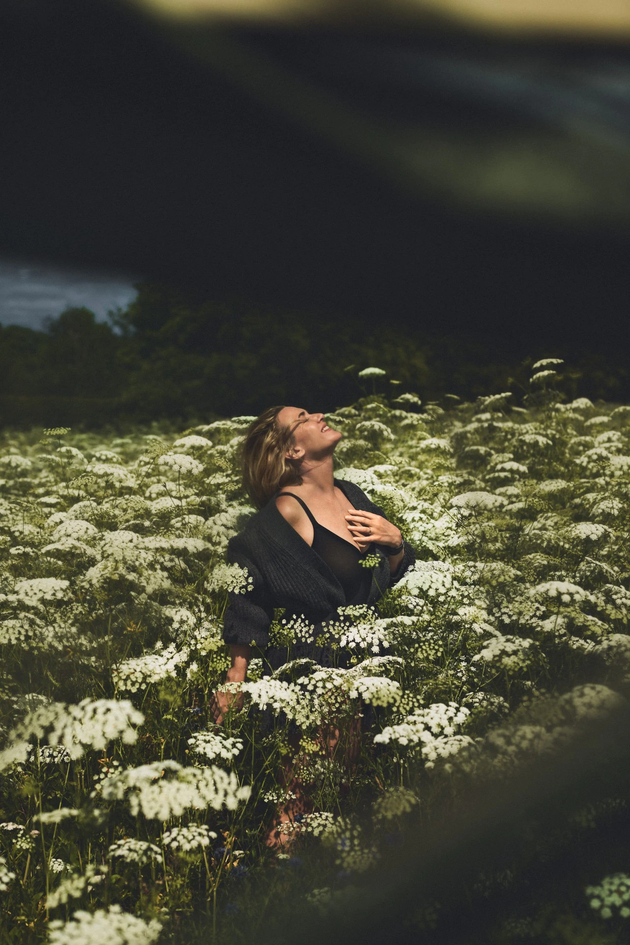 Woman stands in a field of white wildflowers, gazing upwards in outdoor portrait showcasing fashion and nature.
