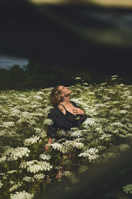 Woman stands in a field of white wildflowers, gazing upwards in outdoor portrait showcasing fashion and nature.