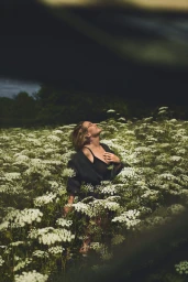 Kate Winslet standing amidst a dense field of white wildflowers wearing a dark textured cardigan over a black top