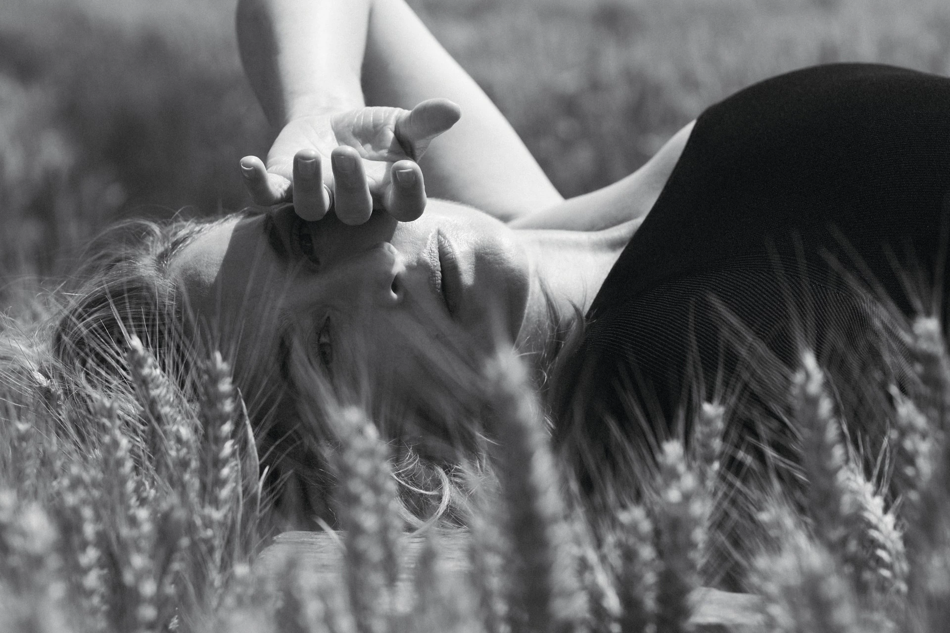 Black and white portrait of a blonde woman lying in a field of wheat, gazing up with her hand above her face in an outdoor setting.