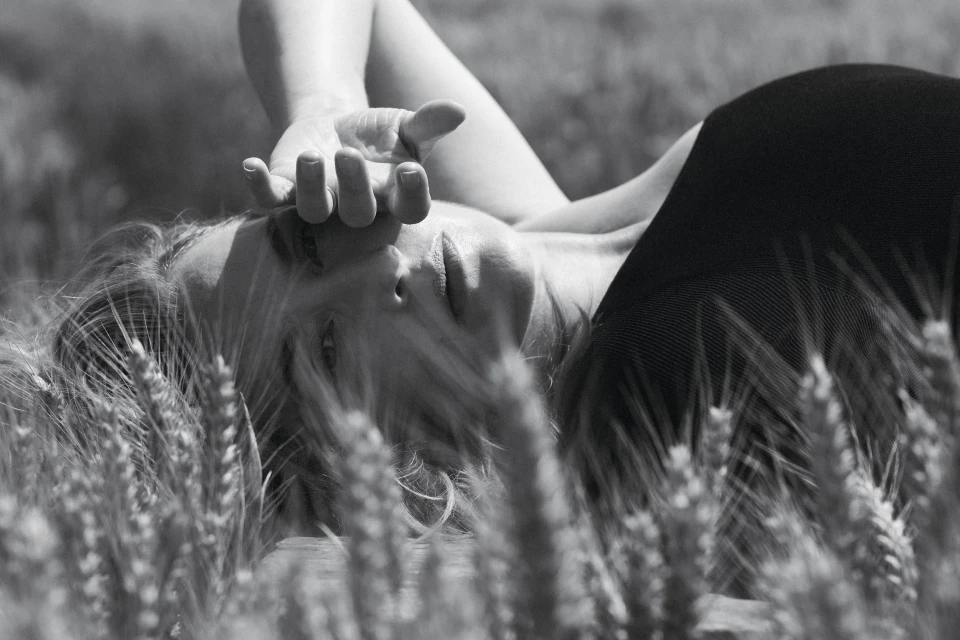 Black and white portrait of a blonde woman lying in a field of wheat, gazing up with her hand above her face in an outdoor setting.