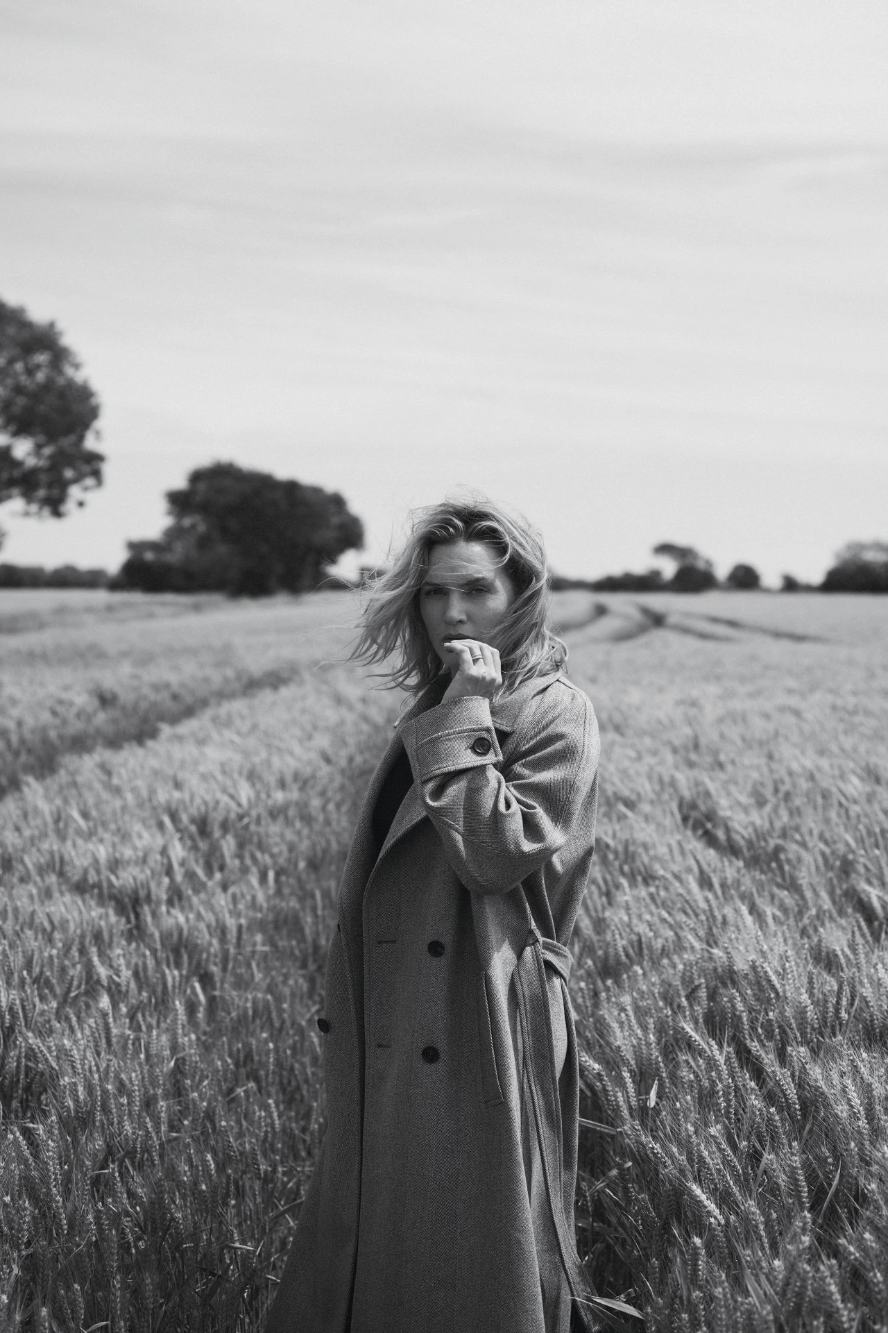 Black and white portrait of a woman in a trench coat standing in a wheat field on a sunny day with clouds in the distance.