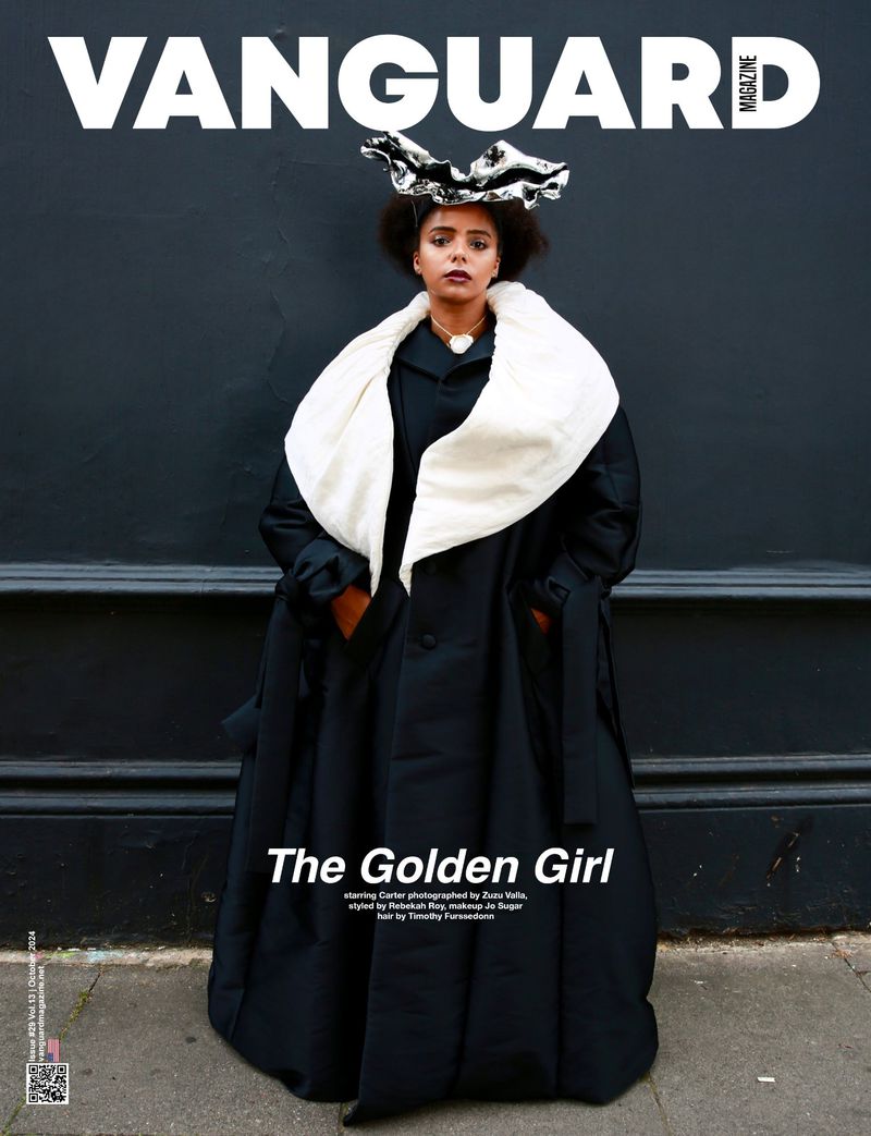Vanguard Magazine cover featuring a woman wearing a black coat with white collar, unique hat, standing against a dark textured wall.
