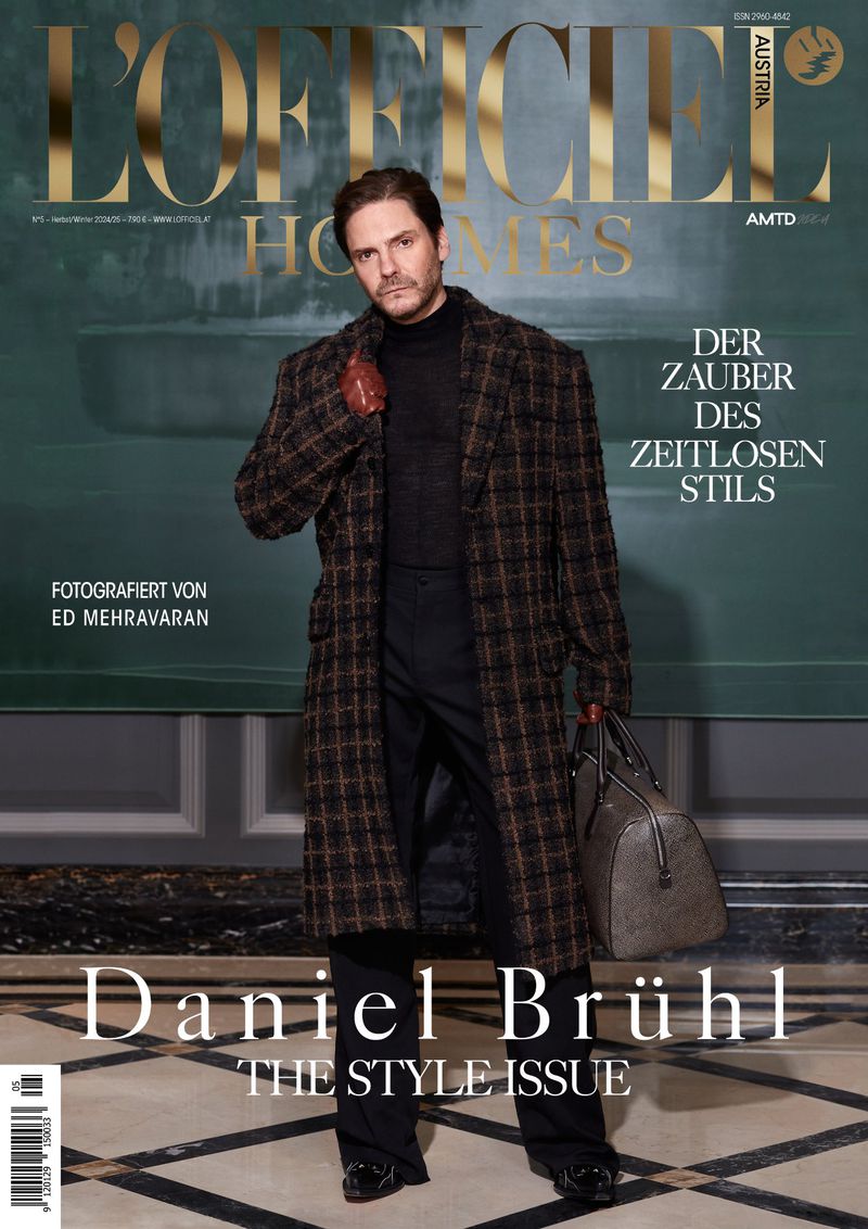 Daniel Brühl on L'Officiel Hommes Austria cover wearing a plaid coat and black turtleneck, holding a duffel bag in an indoor setting.