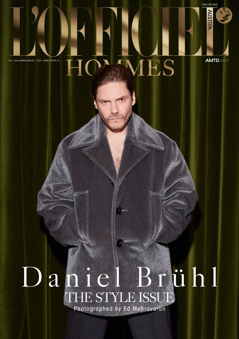 Magazine cover featuring Daniel Brühl in a grey jacket, styled for L'Officiel Hommes, with a green velvet curtain backdrop and clean studio lighting.