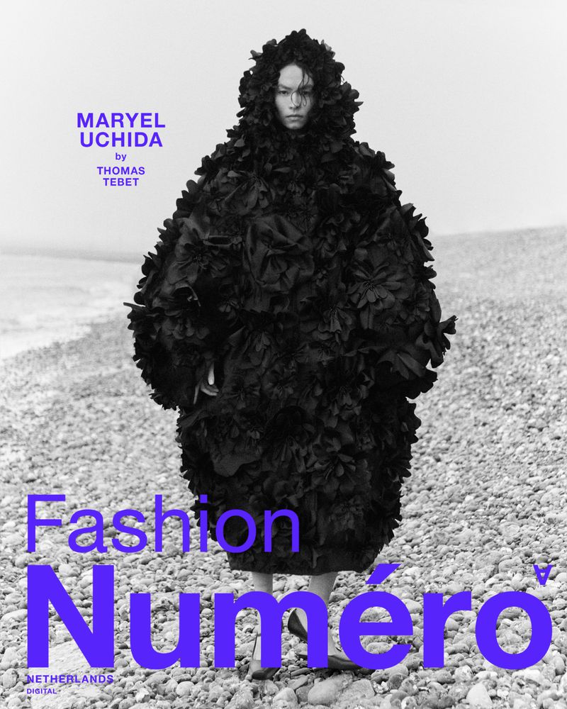 Fashion editorial shot of a person in a large, textured black hooded cloak standing on a rocky beach for Numero Netherlands magazine.