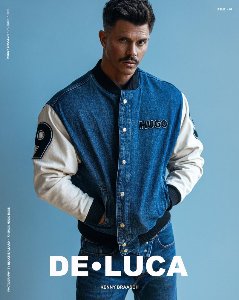 Studio shot of a man with dark hair and a mustache wearing a blue denim jacket with white sleeves, jeans, and a serious facial expression.