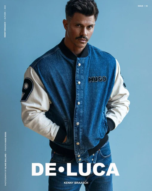 Studio shot of a man with dark hair and a mustache wearing a blue denim jacket with white sleeves, jeans, and a serious facial expression.