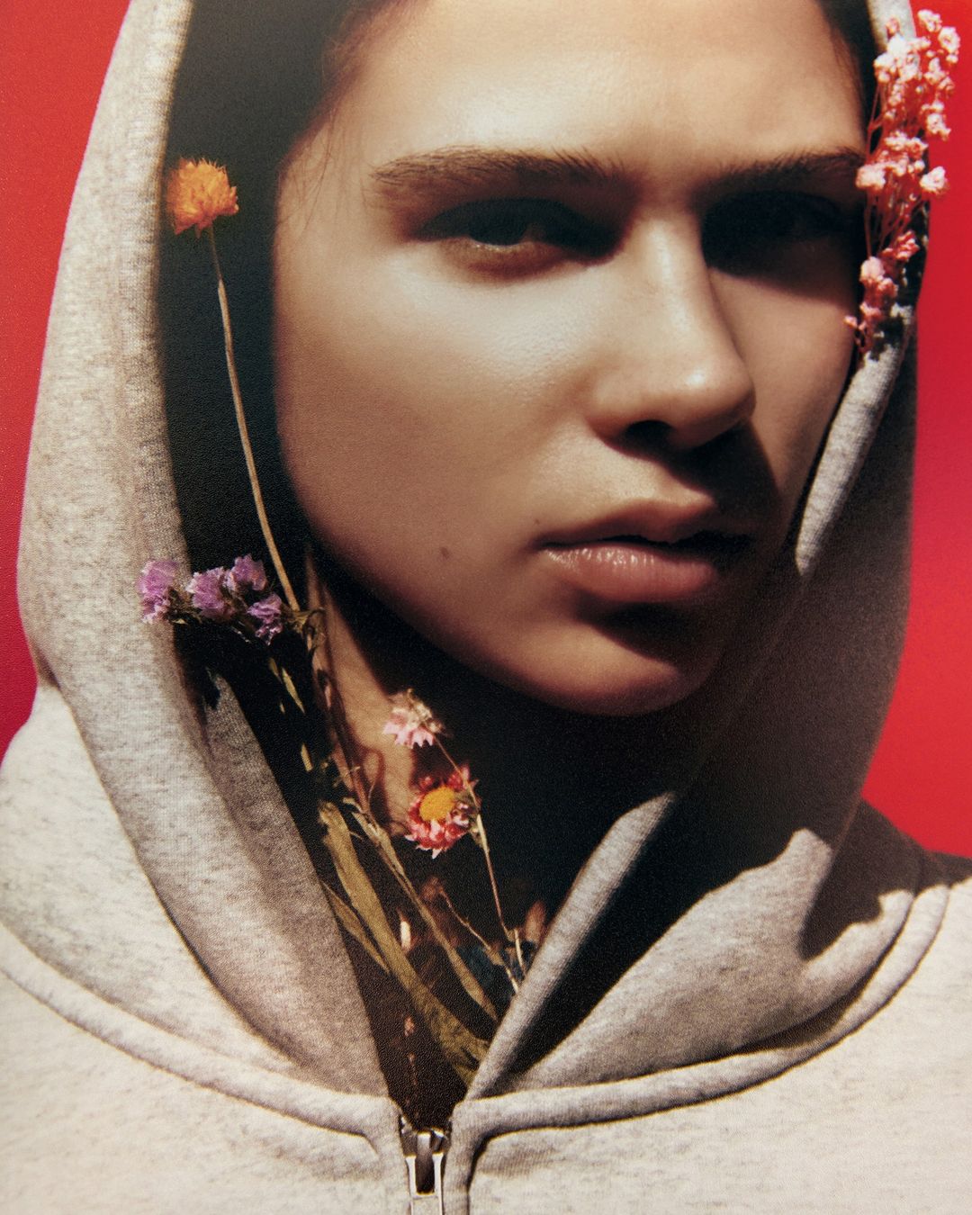 Close-up studio portrait of a woman with fair skin and a neutral expression, wearing a grey hooded sweatshirt and adorned with small flowers.