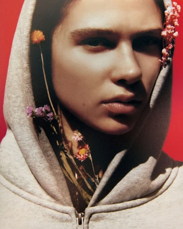 Close-up studio portrait of a woman with fair skin and a neutral expression, wearing a grey hooded sweatshirt and adorned with small flowers.