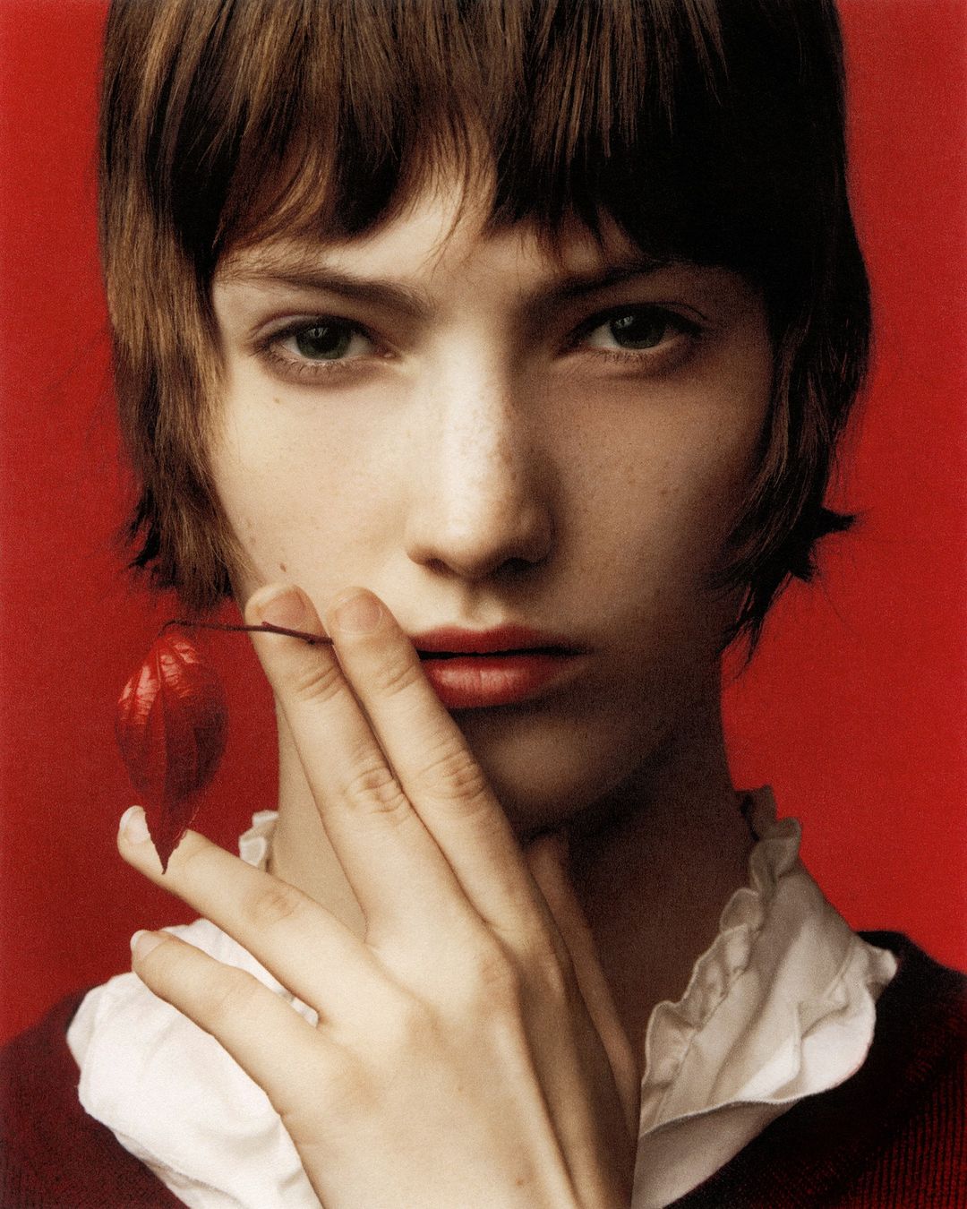Portrait of a brunette female model with her hand near her face, holding a red chinese lantern plant against a red studio backdrop.